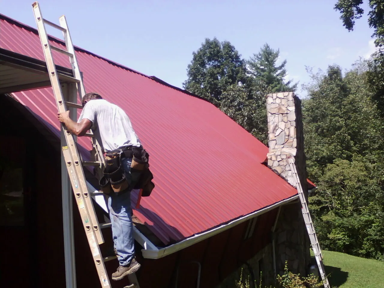 Roofer on a ladder beside a red metal roof and stone chimney — Soffit & Fascia Repair by Roofing Brothers in Robesonia